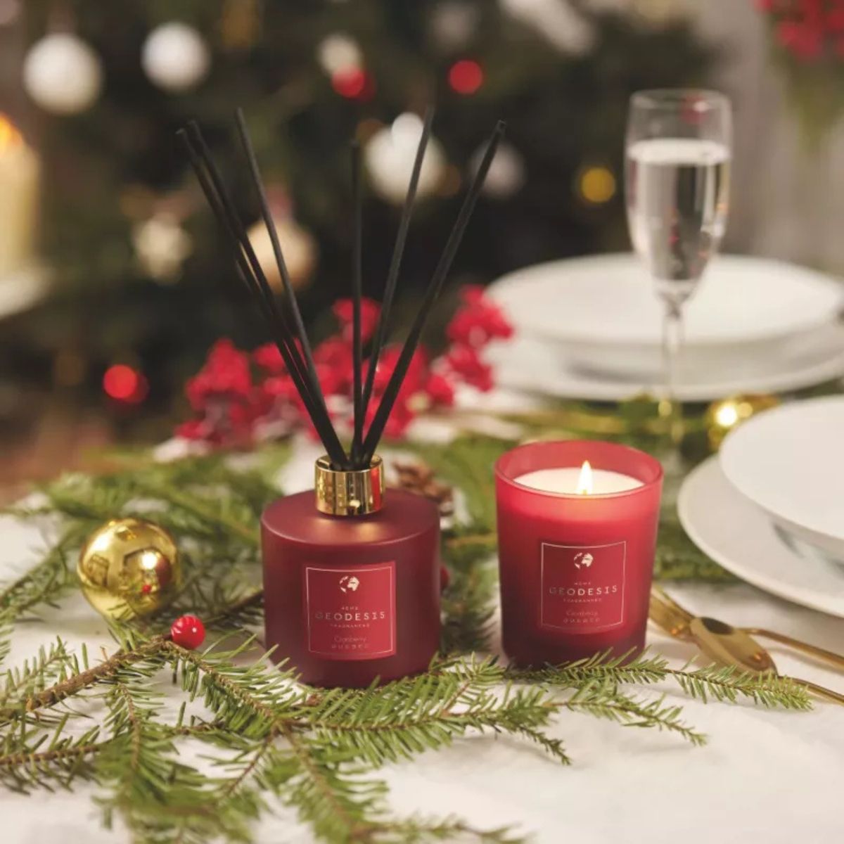Geodesis Cranberry – diffusore con bastoncini e candela profumata rossa, tra rami di abete e decorazioni di Natale.